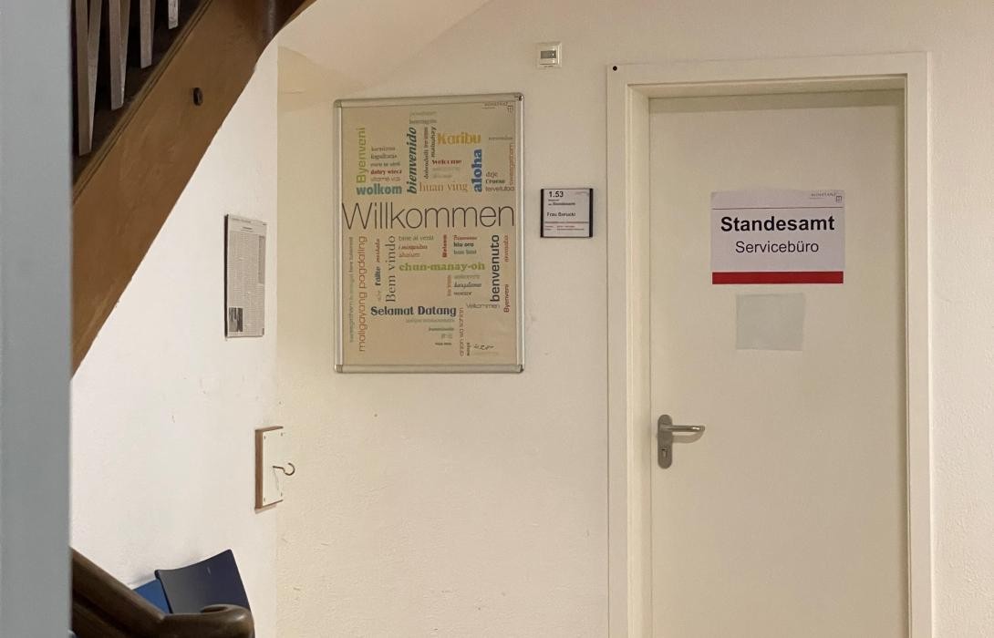 Blick in einen Vorraum, an einer Tür hängt ein Schild mit Info Standesamt  Servicebüro. Daneben ein buntes Poster mit dem Wort Willkommen in verschiendenen Sprachen,