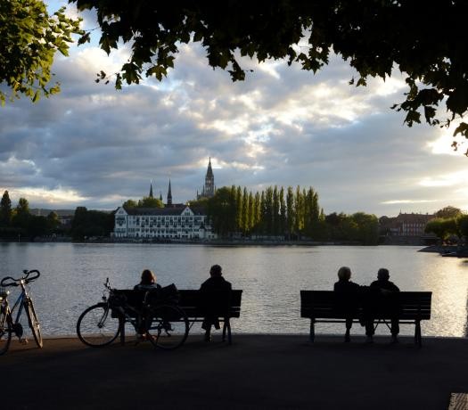 Sitzende Personen auf zwei Bänken am Seeufer; im Hintergrund die Konstanzer Altstadt mit dem Inselhotel