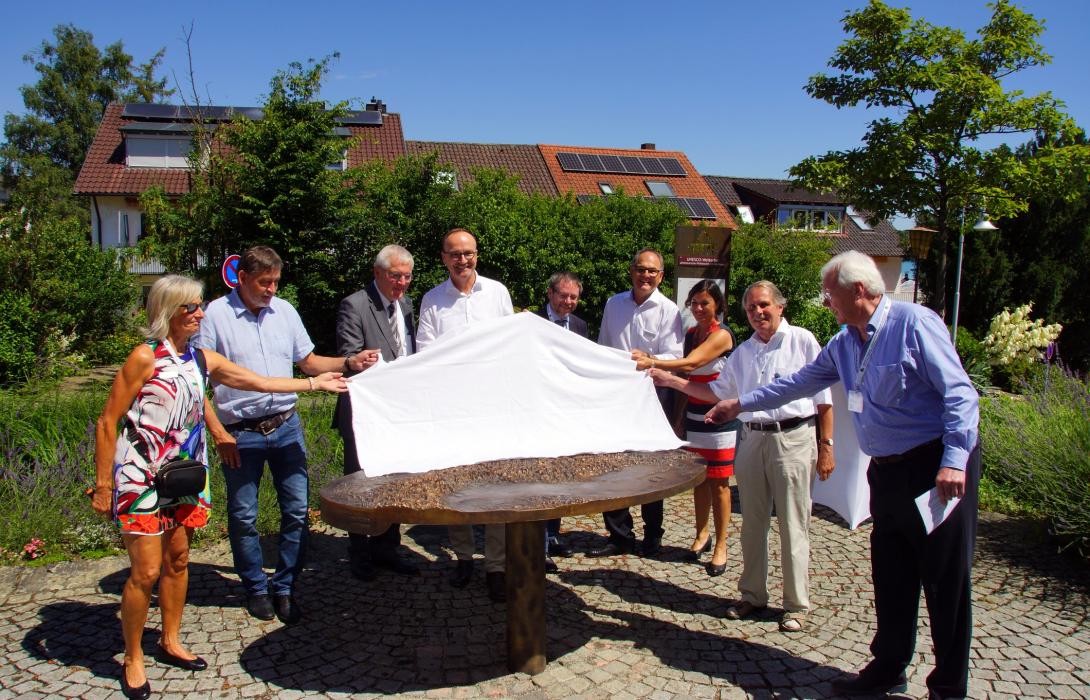 Eine Gruppe Menschen zeiht ein weißes Tischtuch von einem Bronzerelief