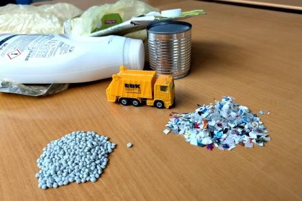 Plastikverpackungen, zerkleinertes Plastik und Granulat liegen in kleinen Haufen auf einem Tisch um ein Spielzeug-EBK-Müllfahrzeug. 