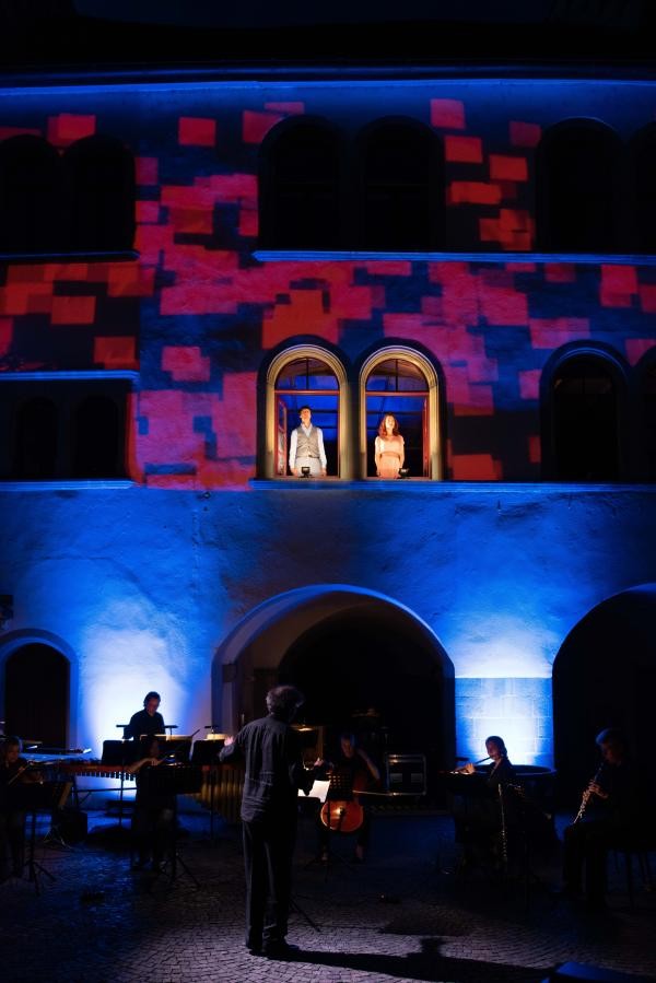Die Fassade des Rathause ist blau-rot beleuchtet, in zwei Fenstern im ersten Stock stehen zwei Personen, davor das Orchester