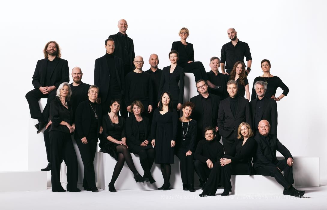 Chorsänger und -sängerinnen in schwarzer Kleidung vor weißem Hintergrund.