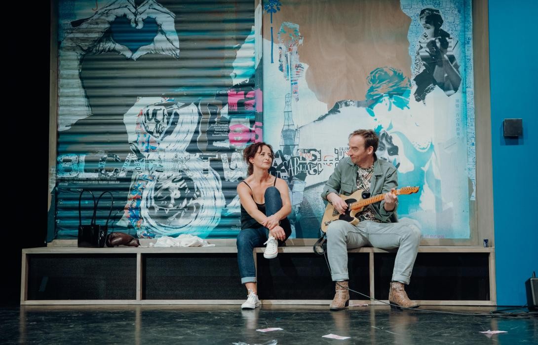 Eine Frau und ein Mann sitzen auf einer Bank, der Mann spielt Gitarre.