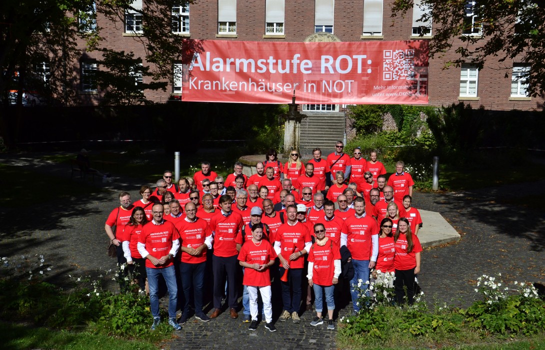 Eine große Personengruppe steht vor einem Gebäude, an dem ein Banner mit der Aufschrift hängt: Alarmstufe Rot: Krankhäuser in Not!" Alle Personen tragen rote T-Shirts.