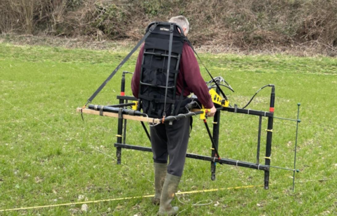 Mann mit einem am Rücken festgeschallten geomagnetischen Vermessungsgerät auf einer Wiese