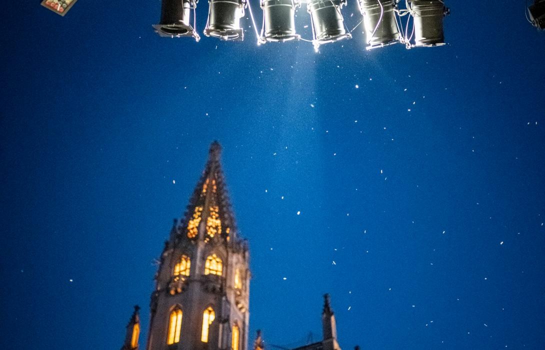 Blick von unten hoch zur beleuchteten Spitze des Münsters, im Hintergrund ist der Abendhimmel zu sehen. Außerdem sieht man von unten einen Stahlträger mit Bühnenlichtern.