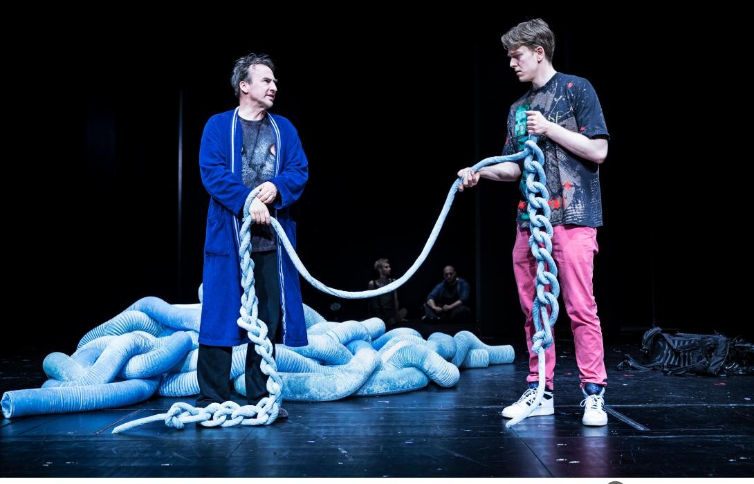 Zwei Personen auf der Bühne die ein Seil, das einen überdimensionalen Wollfaden darstellen soll, zwischen sich halten.