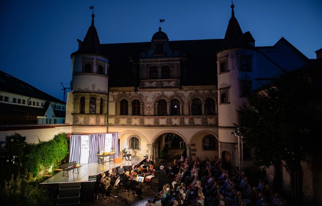 Blick in einen Hof mit Bühne und Publikum in Stuhlreihen und Orchester davor. Es ist bereits dunkel, im Hintergrund sieht man ein mittelalterliches Gebäude mit Rundbogenfenstern und zwei Türmchen. 