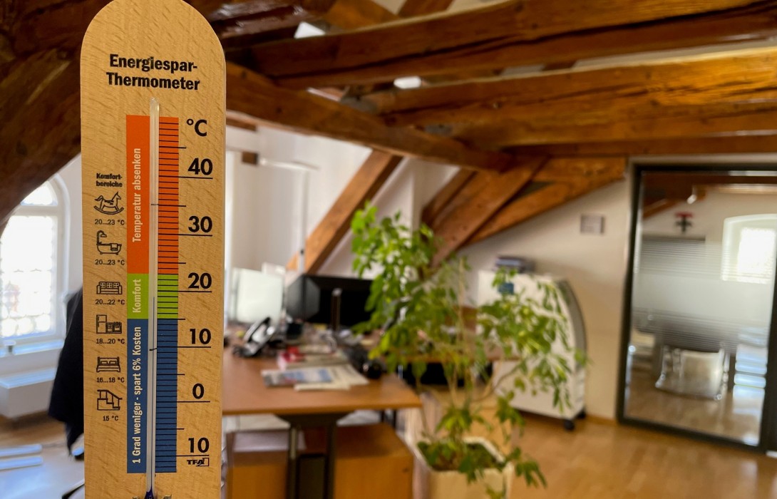 im Vordergrund ein hölzernes Thermometer, das 19 Grad Celsius anzeigt; im Hintergrund ein Büroraum