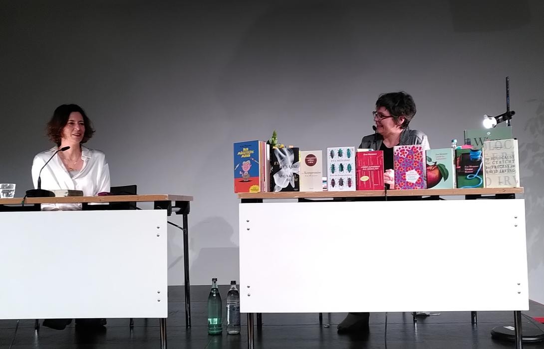 Zwei Frauen sitzen an je einem Tisch, darauf Mikrophone. Auf einem Tisch stehen Bücher aufgereiht.