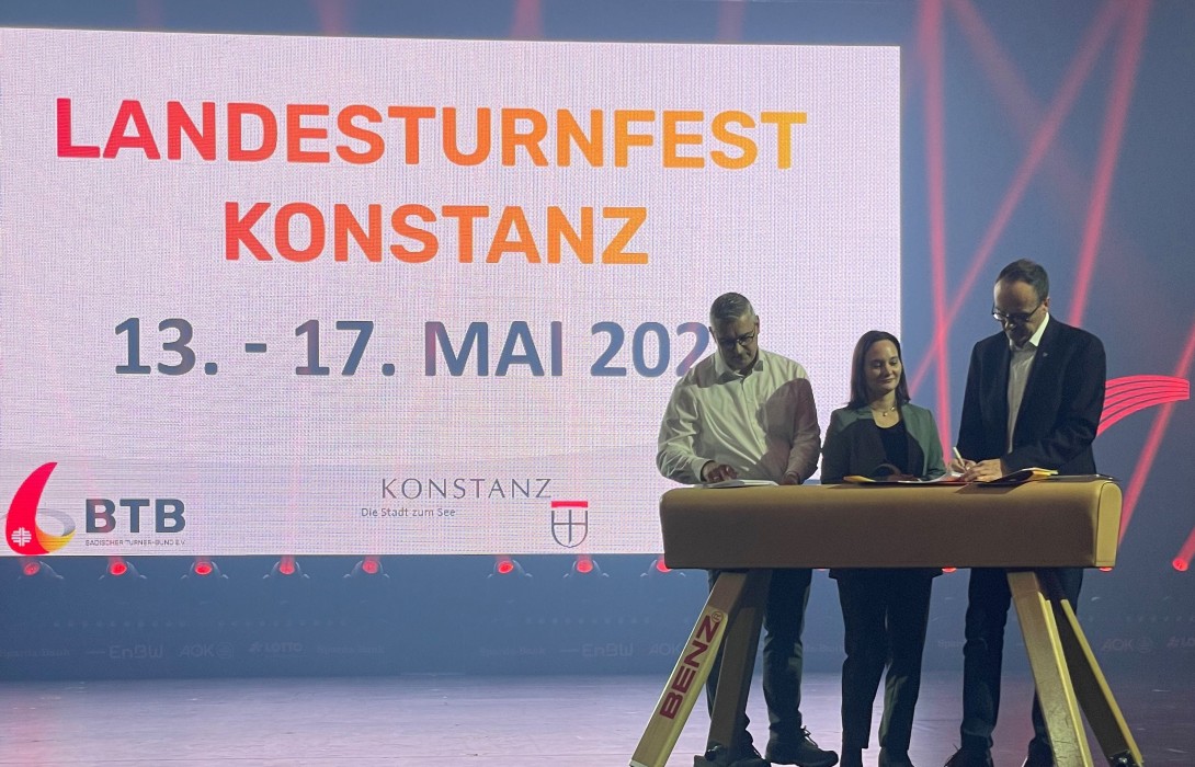Drei personen stehen an eine Turn-Pferd und schreiben. Im HIntergrund sieht man eine Projektion mit Text: "Landesturnfest Konstanz 13. - 17. Mai 2026"