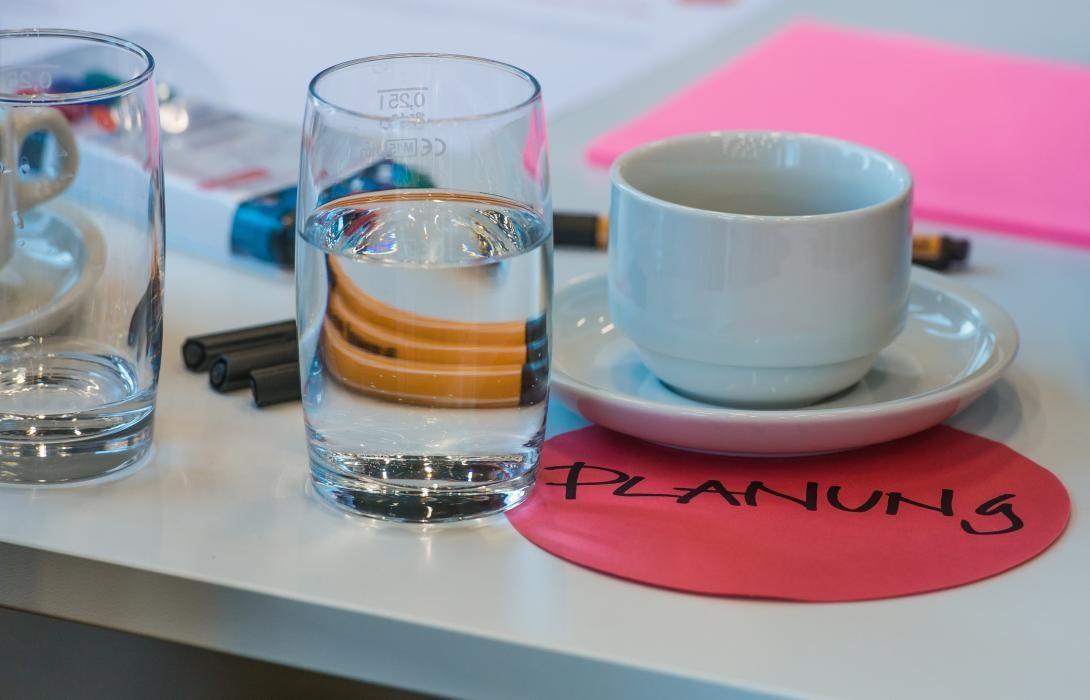 Blick auf einen Tisch: eine Tasse, zwei Gläser, Stifte und buntes Papier. Auf einem Papier ist der Begriff Planung notiert.