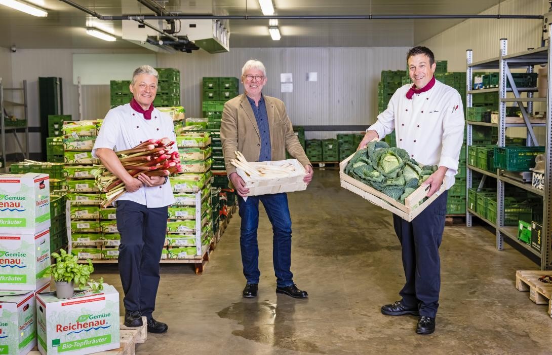 Drei Männer stehen in einem Lagerraum und halten Kisten mit Gemüse in die Kamera