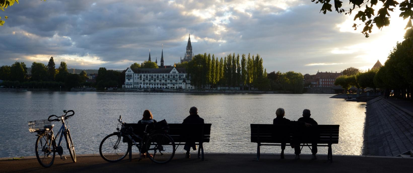 Sitzende Personen auf zwei Bänken am Seeufer; im Hintergrund die Konstanzer Altstadt mit dem Inselhotel