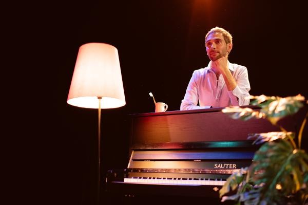 Ein Mann lehnt von hinten auf einem Klavier, man sieht die Tasten, links daneben steht eine Stehlampe und am rechten Bildrand eine Grünpflanze
