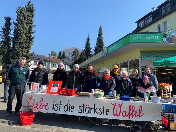 Ein Gruppe Menschen vor dem Geschäft "Konstanzer Landmarkt" steht hinter einem Tisch auf dem "Liebe ist die stärkste Waffel" steht