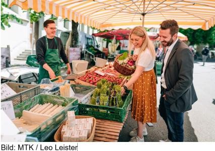 Wochenmarktstand in Konstanz mit Verkäufer, Kundin und Kunde. Als Warenangebot sind grüner und weißer Spargel und im Hintergrund Erdbeeren zu sehen. Die Kundin begutachtet einen Bund grünen Spargels und hat einen Korb mit Gemüse im Arm.