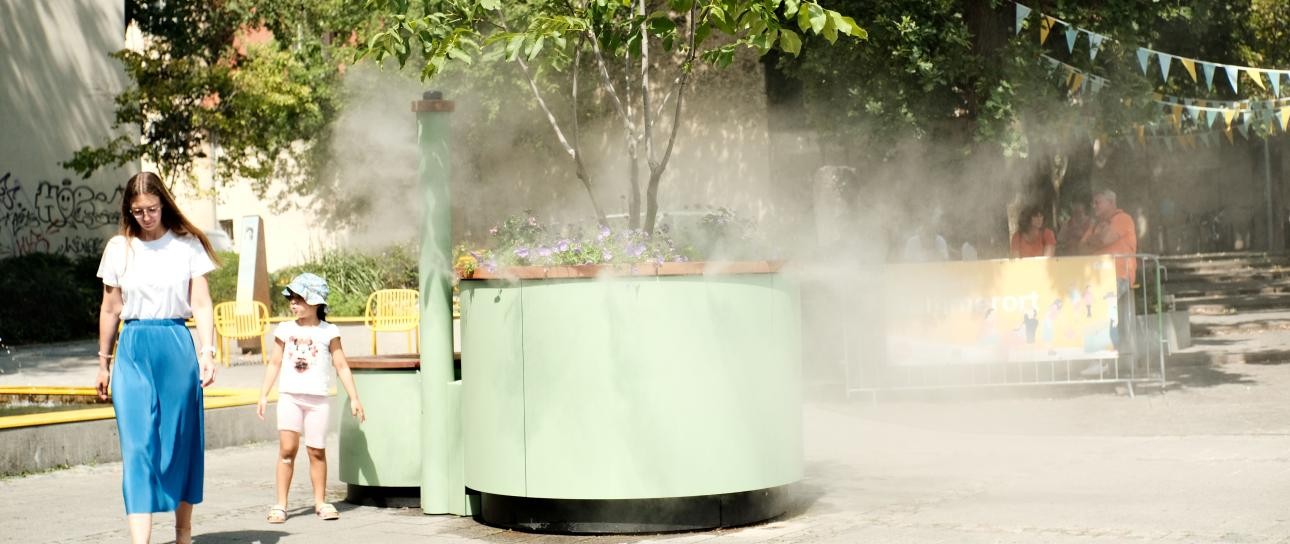 Menschen sitzen am Wasser; drumherum eine parkähnliche Anlage