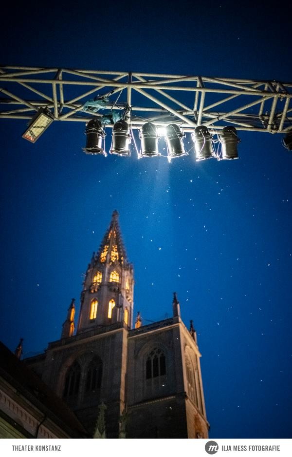 Blick von unten hoch zur beleuchteten Spitze des Münsters, im Hintergrund ist der Abendhimmel zu sehen. Außerdem sieht man von unten einen Stahlträger mit Bühnenlichtern.