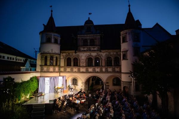 Blick in einen Hof mit Bühne und Publikum in Stuhlreihen und Orchester davor. Es ist bereits dunkel, im Hintergrund sieht man ein mittelalterliches Gebäude mit Rundbogenfenstern und zwei Türmchen. 
