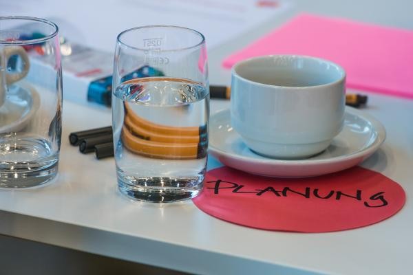 Blick auf einen Tisch: eine Tasse, zwei Gläser, Stifte und buntes Papier. Auf einem Papier ist der Begriff Planung notiert.