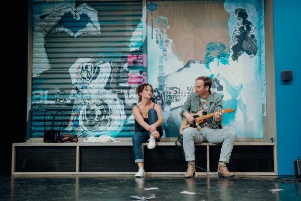 Eine Frau und ein Mann sitzen vor einer Wand mit Graffiti, der Mann spielt auf einer E-Gitarre und schaut die frau an. Sie hat ein Knie angewinkelt und schaut den Mann an.