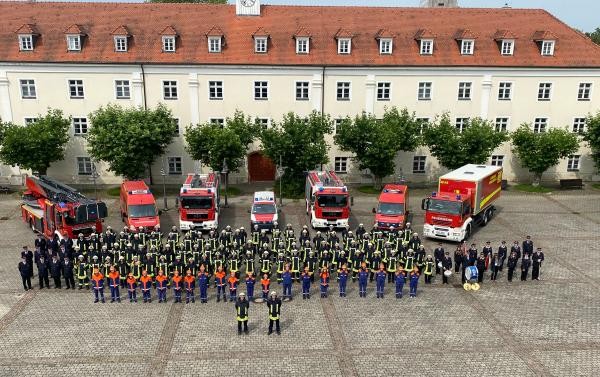 Luftbild, man sieht ein große Gruppe Menschen in Uniform vor 7 unterschiedlichen Feuerwehrautos stehen.
