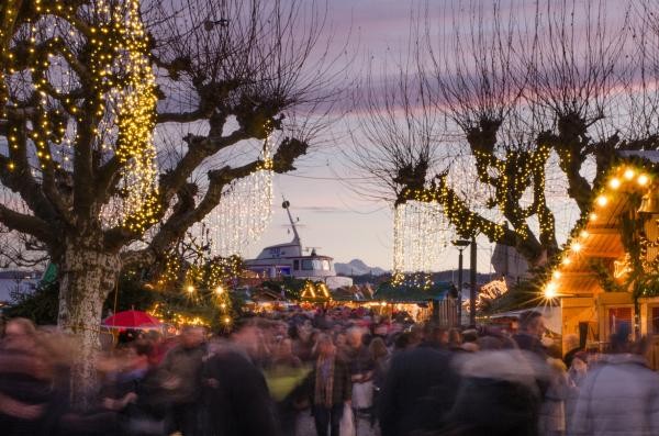 Blick über die Köpfe der BesucherInnen eines Weihnachtsmarkts. Im Hintergrund mit Lichterketten behängte Bäume und Buden