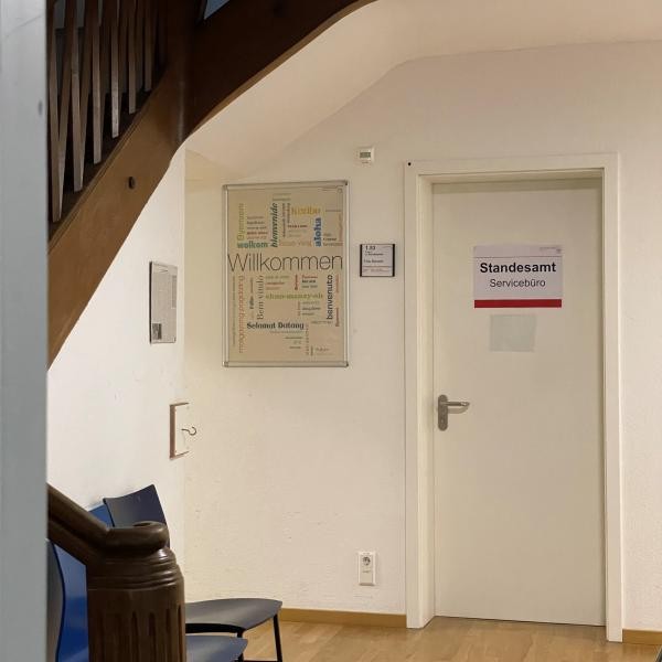 Blick in einen Vorraum, an einer Tür hängt ein Schild mit Info Standesamt  Servicebüro. Daneben ein buntes Poster mit dem Wort Willkommen in verschiendenen Sprachen,