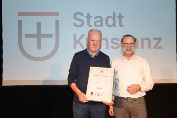Zwei Personen stehen auf einer Bühne, im Hintergrund ist das Logo der Stadt Konstanz zu sehen, die Person links hält eine Urkunde.