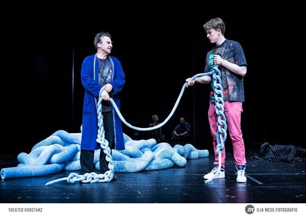 Zwei Personen auf der Bühne die ein Seil, das einen überdimensionalen Wollfaden darstellen soll, zwischen sich halten.