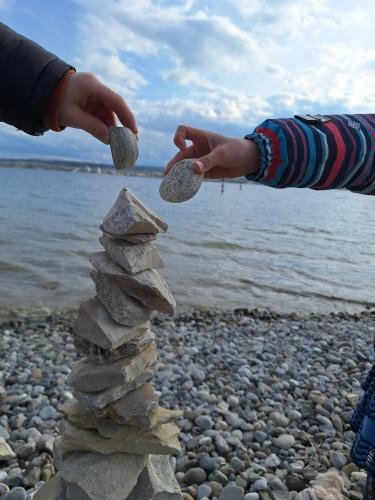 Ein Turm aus steinen am Bodenseeufer, zweit Kinderhände legen jeweils noch einen weitern Steinoben drauf