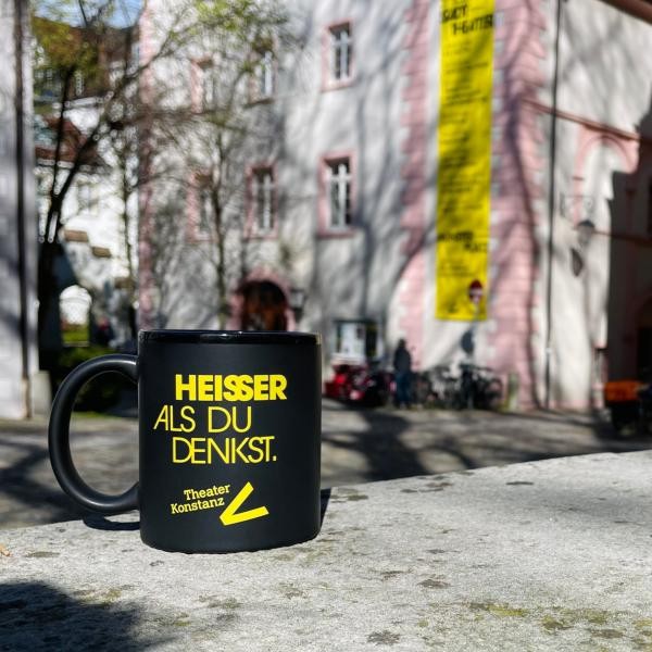 Eine schwarze Tasse mit gelber Schrift "Heisser als du denkst", im Hintergrund sieht man das Theatergebäude