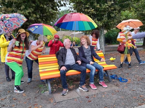Bunt gestrichene Bank, rundherum einige bunt gekleidete Menschen stehend und sitzend, mit Regenschrimen