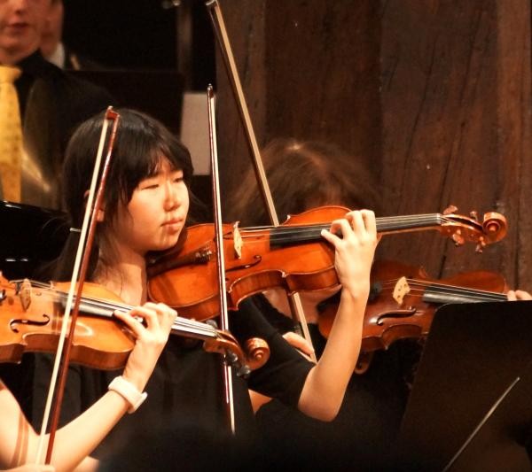 Eine junge Frau mit schwarzen Haaren und schwarzem Kleid hält eine Violine in der Hand. Sie steht in einem Orchester.