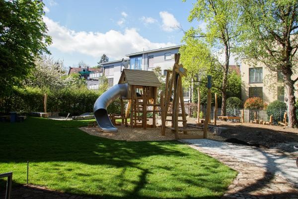 Ein Klettergerüst aus Holz mit Rutsche aus glänzendem grauem Aluminium steht mitten in einem Spielplatz umgeben von grünem Rasen.