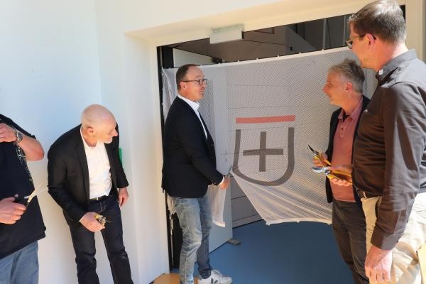 Eine Gruppe Männer steht links und rechts um eine geöffnete Tür. Im Türrahmen hängt eine leicht transparente Flagge mit dem Wappen der Stadt Konstanz. Die zweite Person von links, Oberbürgermeister Uli Burchardt, schneidet die Flagge mit einer Schere durch.