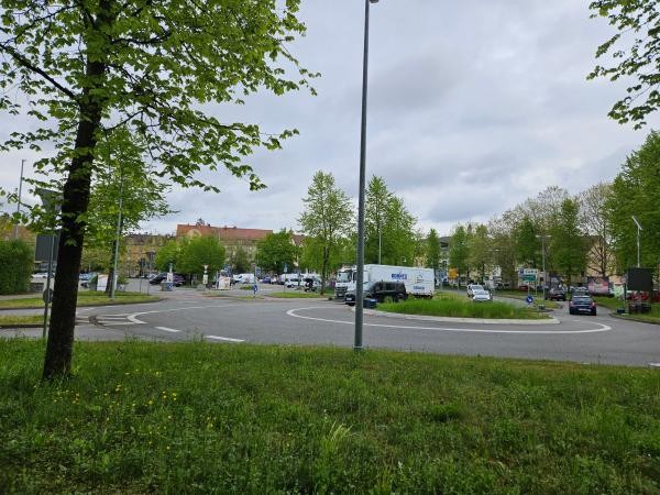 Auf dem Bild ist im Vordergrund eine grüne Wiese zu sehen. Am linken Bildrand befindet sich ein Baum. Im weiteren Verlauf des Bildes ist dann ein begrünter Kreisverkehr zu sehen, der von mehreren Autos umrundet wird.