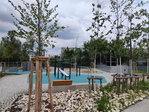 Das Bild zeigt eine moderne Spiel- und Bewegungsfläche in der Grünspange Weiherhof in Konstanz. Zu sehen sind Calisthenics-Geräte auf einem blauen Fallschutzboden, junge Bäume mit Holzstützen, Natursteinflächen und ein Streetballplatz im Hintergrund. Die Anlage ist von Grün umgeben.