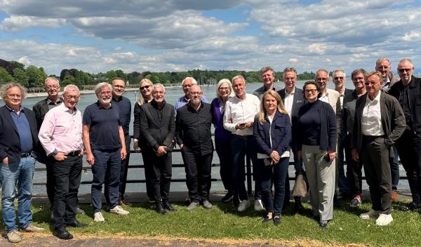 Rund 20 Personen stehen auf einer Wiese, im Hintergrund ein See und bewölkter Himmel
