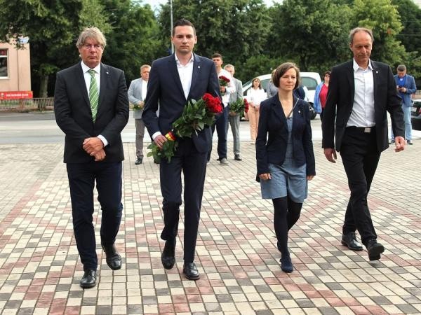 Drei Männer und eine Frau in formeller Kleidung gehen über eine gepflasterte Straße. Der zweite Mann von links grägt einen Strauß roter Rosen mit schwarz-rot-goldener-Binde vor dem m Bauch.