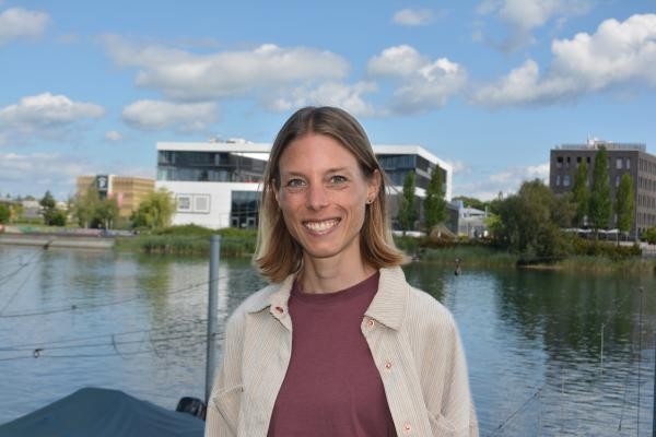 Lächelnde Frau mit dunkelblonden schulterlangen Haaren vor einem Fluss; im Hintergrund Gebäude