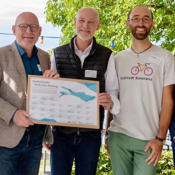 Drei Männer stehen nebeneinander und die zwei links halten ein gerahmtes Zertifikat mit Unterschriften und einer Grafik des Bodensees in der Hand