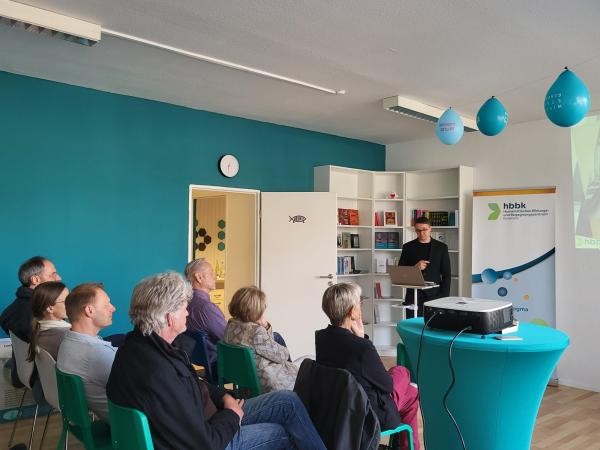 Copyright Stadt Konstanz Am rechten unteren Bildrand sitzen Personen auf Stühlen und hören einem Vortragenden zu. Die Personen sitzen in einem Raum mit türkisfarbenen Wänden.