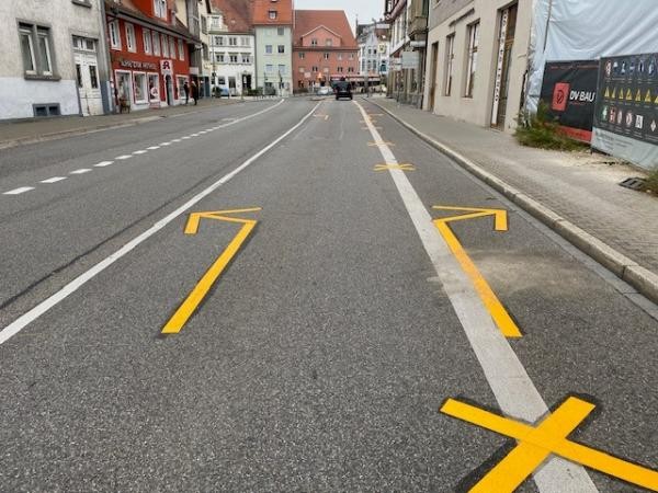 Das Bild zeigt eine Straße in einer Stadt mit frisch aufgebrachten gelben Fahrbahnmarkierungen. Zu sehen sind große Pfeile und Kreuze auf dem Asphalt, die den Verkehr leiten sollen. Entlang der Straße stehen mehrstöckige Gebäude mit Geschäften im Erdgeschoss.