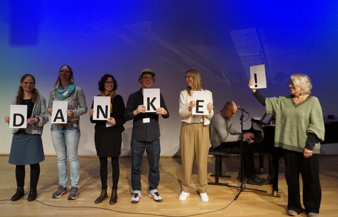 Eine Gruppe Personen, die je einen Buchstben des Wortes "Danke" halten. Im Hintergrund sitzt jemand an einem Klavier.