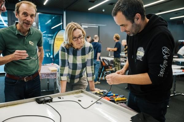 Drei Personen stehen in einem modernen, hell beleuchteten Raum an einem Arbeitstisch und arbeiten gemeinsam an einem Solarmodul. Eine Person rechts hält ein Kabel und greift zu einer Crimpzange, auf dem Tisch liegen weitere Werkzeuge.
