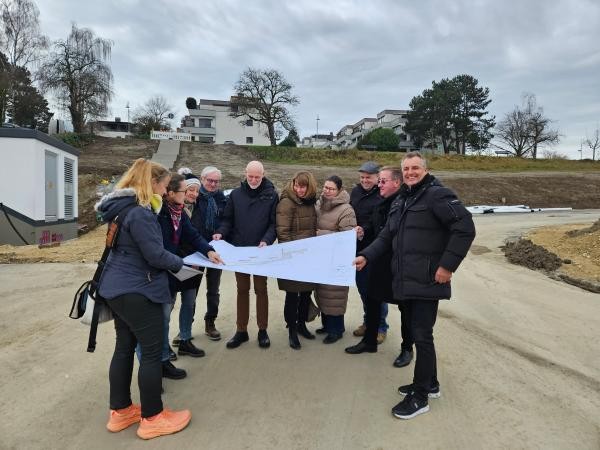 Mehrere Personen stehen auf einer Baustraße im Halbkreis um einen Bauplan herum. Im Hintergrund zu sehen ist Wiese, Bäume und Häsuer.