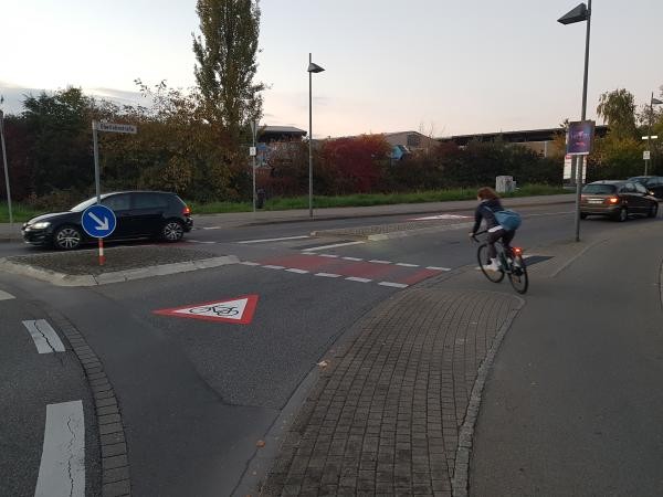 Auf dem Bild zu sehen ist eine Ausfahrt aus einem Kreisverkehr, eine Radfahrerin kreuzt gerade die Straße. Auf der Straße sind in roter Farbe Markierungen aufgebracht, die zu mehr Sicherheit führen sollen.