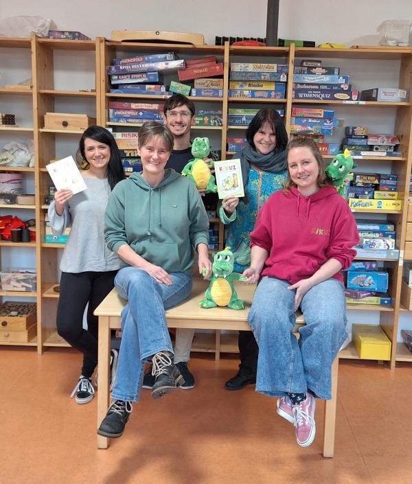 5 Personen auf dem Bild vor einem Regal in dem sich Spiele befinden. zwei Frauen sitzen vorne auf einem Tisch und im Hintergrund stehen zwei Frauen und ein Mann. Es sind Kuscheltierdrachen zu sehen.
